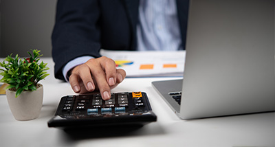 a person using a calculator with one hand while working on a laptop on a desk with a small plant and documents calculating 13 percent