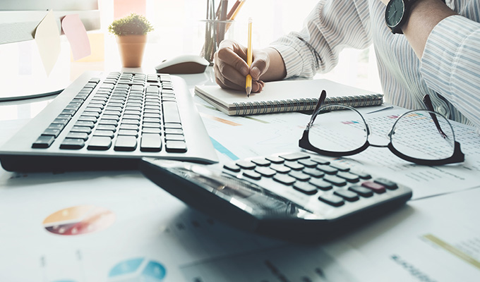 person writing notes with a pencil while using a keyboard calculator and glasses on a business desk with papers and graphs related to 7 strategies for success