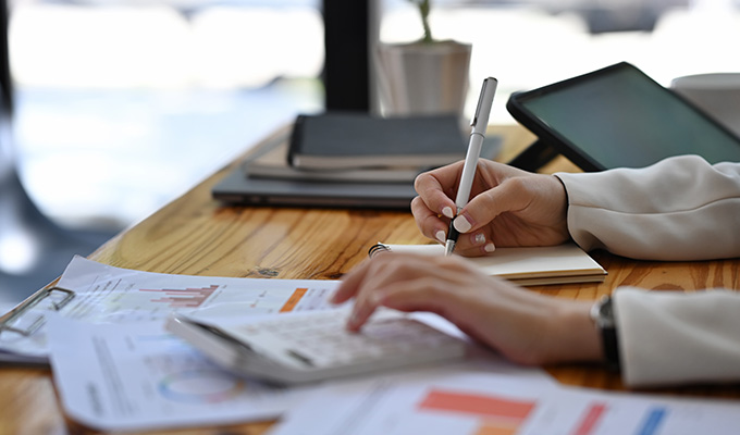 person writing in notepad with calculator and business documents on wooden desk indicating 3 financial points